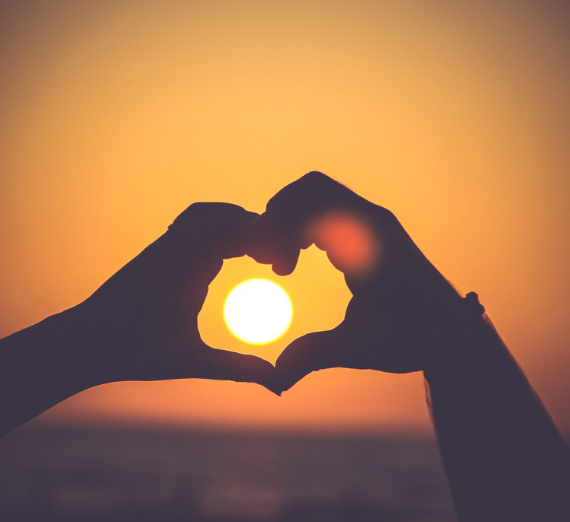 Two hands make a heart shape and frame the sun during a sunset