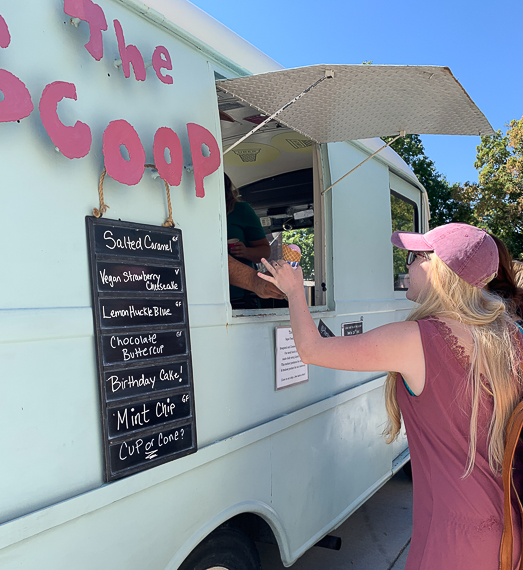 A student reaches up to take her ice cream cone from the window of The Scoop ice cream truck. Written on the side of the van is "The Scoop" and the list of flavors.