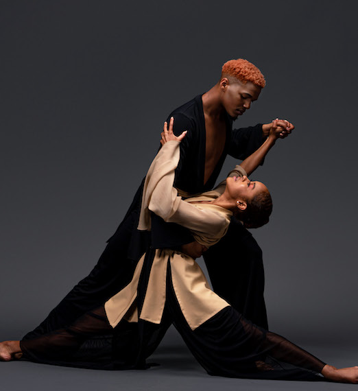 Two people of African American heritage dancing. One is male with short red hair, wearing black. The other is female with a khaki robe.