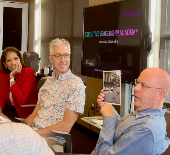 Man with glasses holding up photo while another man with glasses and woman look toward him while smiling.