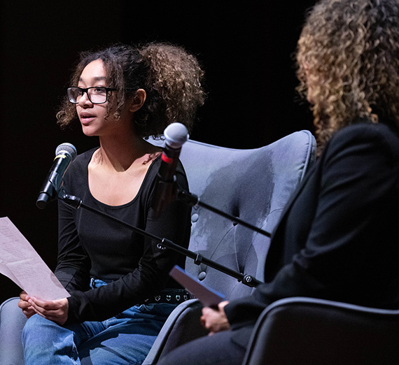 A young black woman sitting on stage with a microphone in front of her and another woman next to her. 