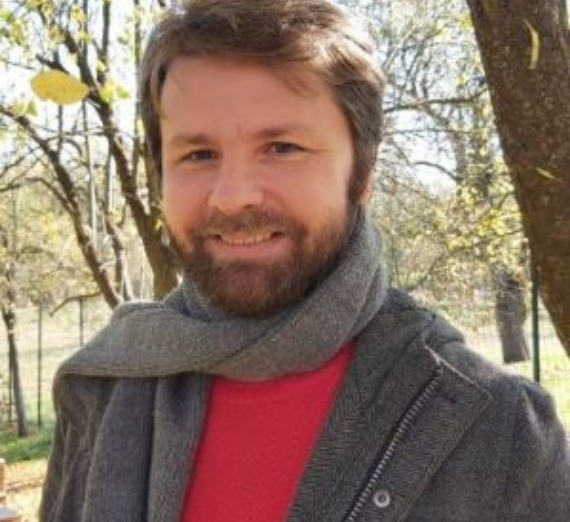 Man with red shirt, grey jacket and grey scarf smiles at camera in front of treed background.