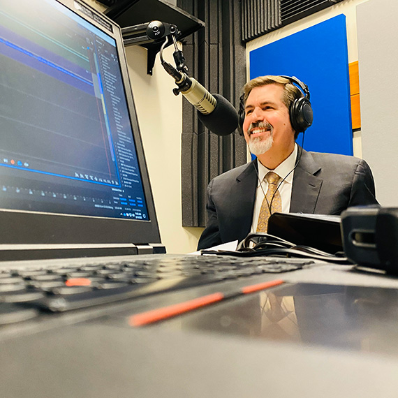 A laptop computer in the foreground and President Thayne McCulloh behind a podcast microphone in the background.