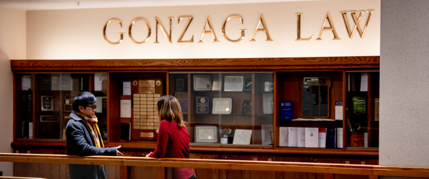 Two students standing at a rail talking in front of a Law School memorabilia cabinet with the words "Gonzaga Law" in gold over the cabinet.