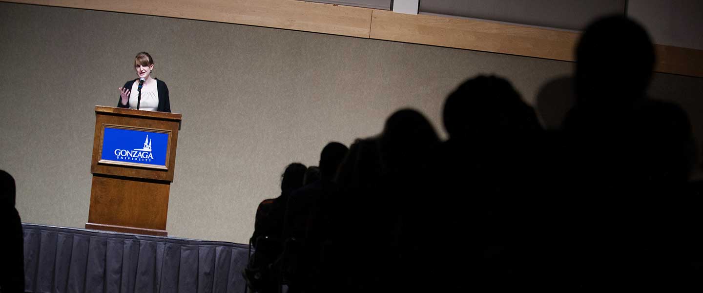 A lecturer stands at a podium on the left with a darkend audience to the right.