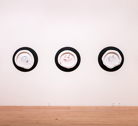 Artwork displayed on the white walls of the Jundt Art Museum. Three, black spheres with a blend of white and red or blue paint in the center.