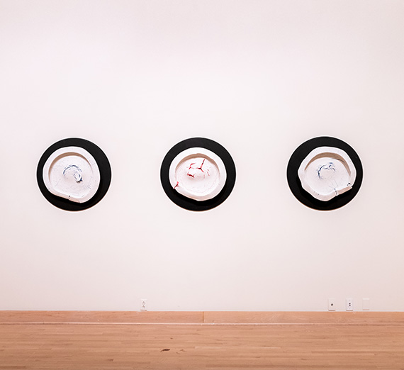 Artwork displayed on the white walls of the Jundt Art Museum. Three, black spheres with a blend of white and red or blue paint in the center. 
