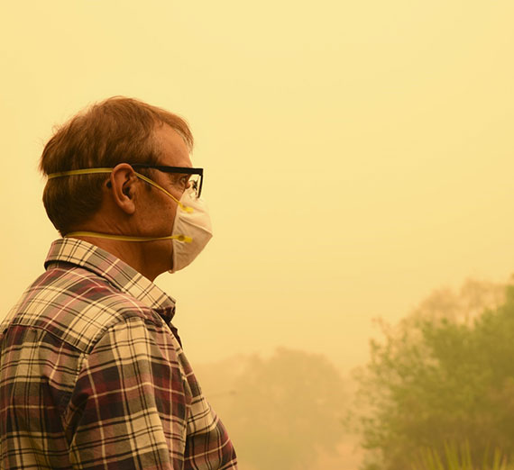 A man wearing glasses and a protective mask stands against a backdrop of dense, yellowish haze.