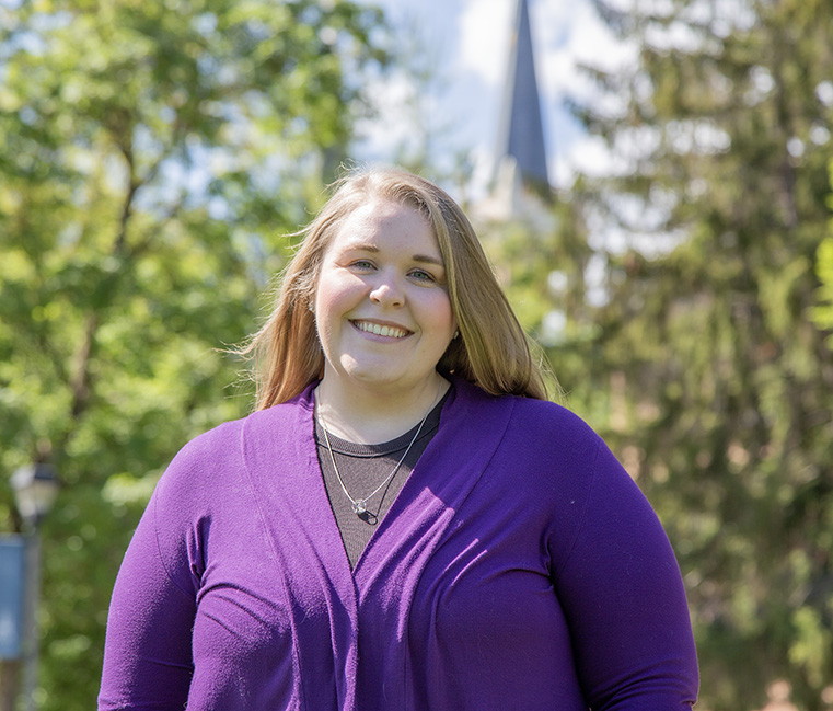 Master's in Communication and Leadership Studies alumna, Rebekah Buettner standing on campus with trees and the spires behind her.