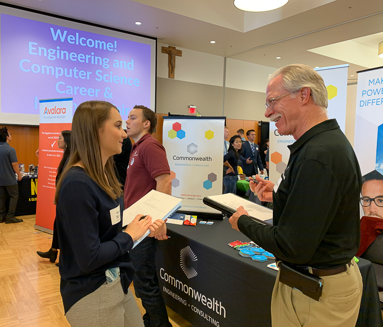 A young woman holds a stack of papers in her arm as she talks with an employer representative at the Engineering and Computer Science Career Fair in 2019.