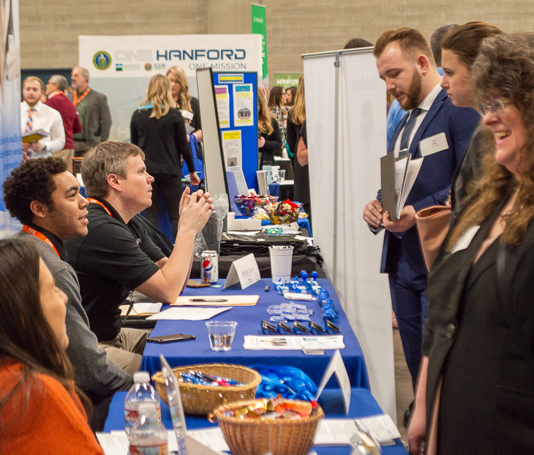 A long table stretching away from the camera shows a row of employer representatives sitting on the left side, with students and alumni standing on the right side. At the furthest end of the table, more students are seen walking past similar employer tables at the FUSE Career Fair in 2019.