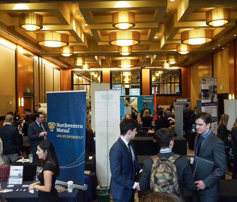 Students in professional clothing mill around employer booths composed of tables with pop-up banners in the Renaissance ballroom in Seattle during the Seattle Trek Career Fair.