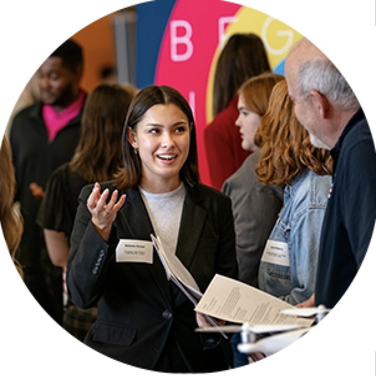 A young woman in professional attire speaks enthusiastically with an older man at a networking event or career fair. She is holding papers and wearing a name tag, surrounded by other attendees in a colorful, busy setting.