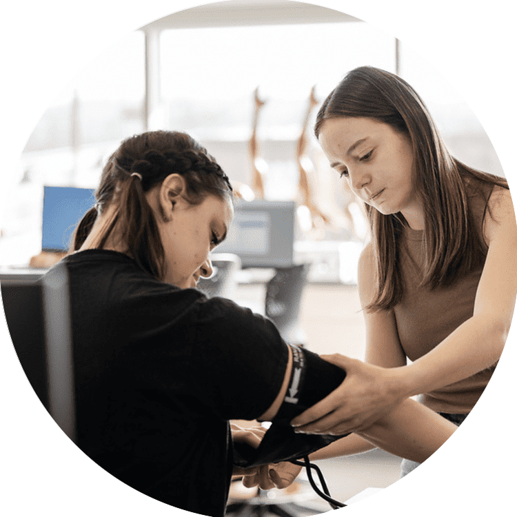 A young woman assists another woman by measuring her blood pressure in a clinical or educational environment. The setting appears bright and modern, with medical or anatomical models visible in the background.