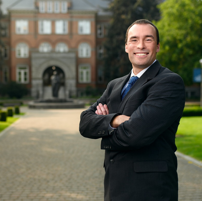 A photo of Steve Keller, Senior Director of Undergraduate Admission. Steve is in the foreground of the photo wearing a black suit with a white shirt and a navy blue tie. He is smiling and crossing his arms over his chest. In the background, is the front of College Hall a large red brick building with a grey stone front on Gonzaga's campus. In front of College Hall is a black statue of St. Ignatius of Loyola.