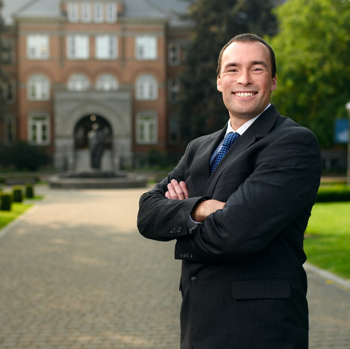 A photo of Steve Keller, Senior Director of Undergraduate Admission. Steve is in the foreground of the photo wearing a black suit with a white shirt and a navy blue tie. He is smiling and crossing his arms over his chest. In the background, is the front of College Hall a large red brick building with a grey stone front on Gonzaga's campus. In front of College Hall is a black statue of St. Ignatius of Loyola. 