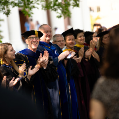 A group of faculty in academic regalia clapping for a procession of students. 