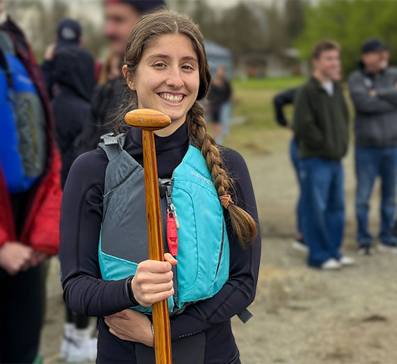 Young woman standing at a lake beach holding an oar and wearing a floatation vest with other young people in the background