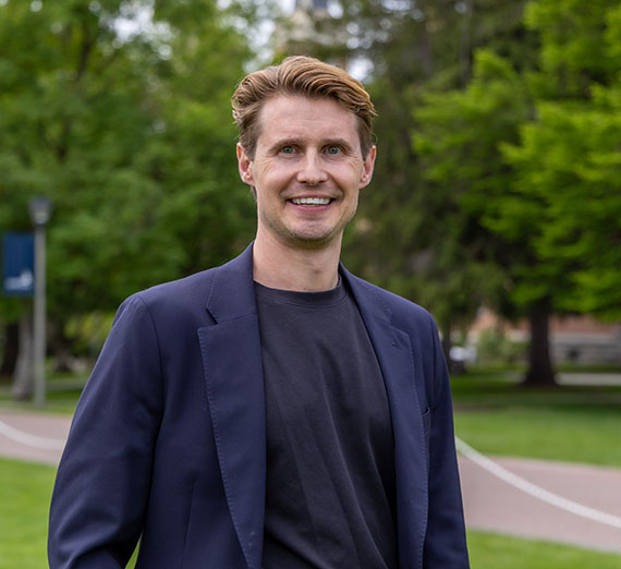 Collin Brum, male wearing navy blazer and black shirt, standing with campus trees and brick walkway in background. 