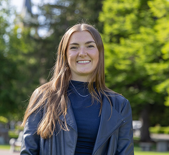Claire Martin, female wearing black shirt with black leather jacket, standing with trees in the background 