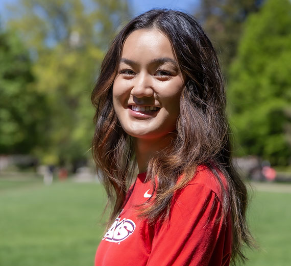 Female student with brown hair and red shirt standing in front of campus greenery 