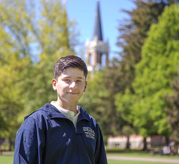 Sebastian Pena wearing blue zip up and standing outside with St. Al's spire in the background behind trees. 