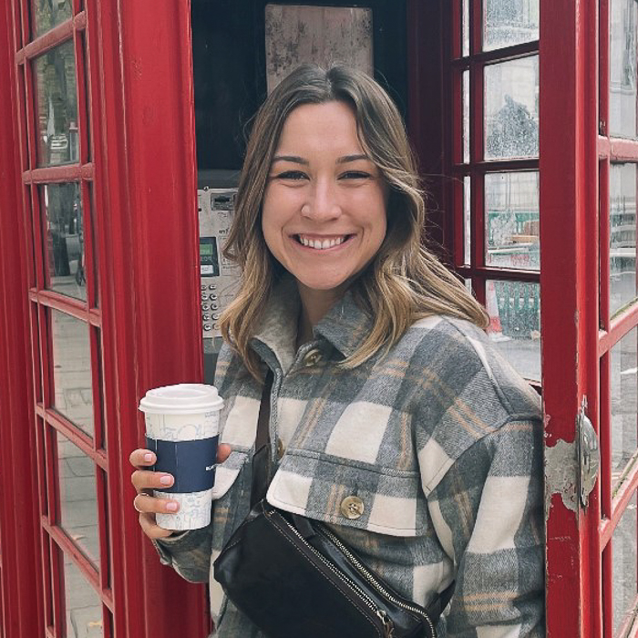 Kelsie exits a red phone booth with a smile, white coffee cup in hand. She wears a plaid shrug over her shoulders. 