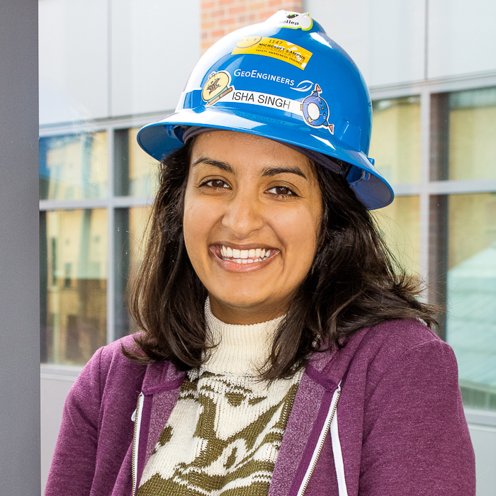 Isha stands in front of the bright glass windows in the Herak walkway wearing a purple sweater and her blue hardhat, which is decorated with stickers. 