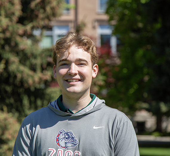 Male student in grey Gonzaga hoodie standing with brick building and trees in background