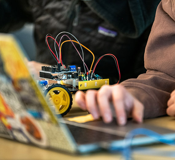 A person typing on a laptop with a robotic car in the background.