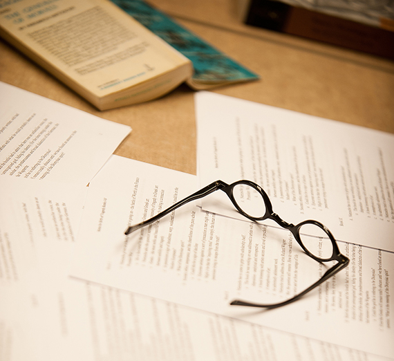 A pair of glasses rests on a pile of papers near an open book on a table