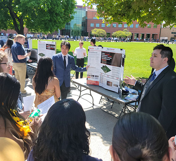 People at tables with academic posters and items edging a lawn outdoors