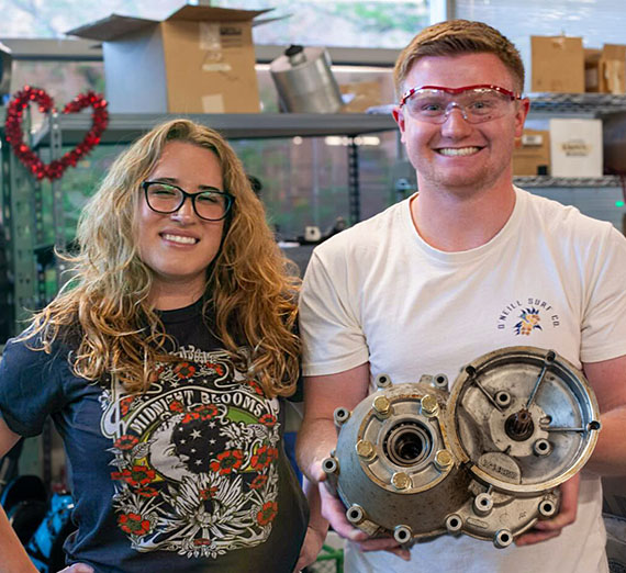 Two students stand in a workshop holding a vehicle transmission.