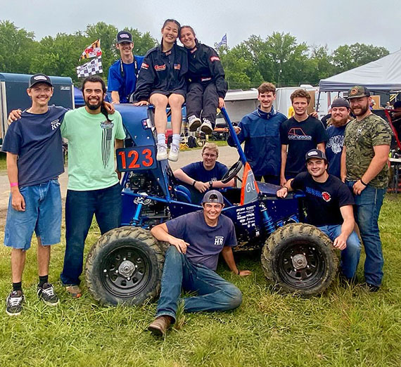 a dozen young adults gathered around a muddy off-road vehicle