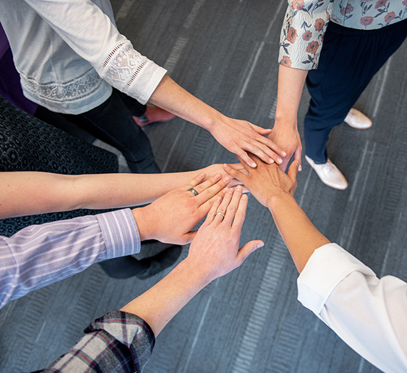 Six people gathered in a circle reaching hands out and connecting in the middle.