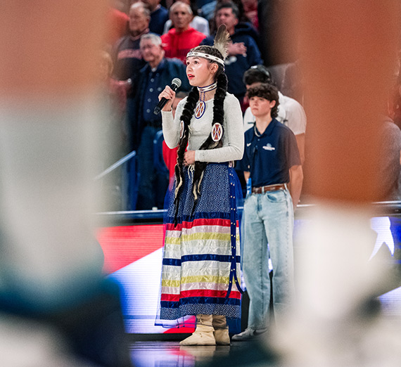 A  Native American woman sings at a N7-sponsored basketball game, which honored Native American heritage and culture.
