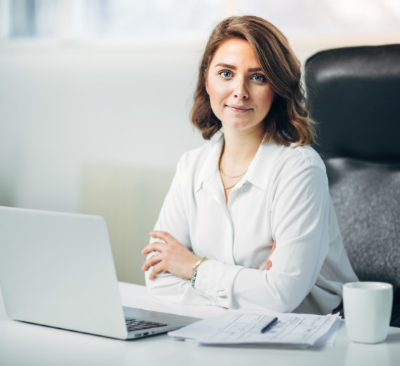 Young woman sitting at laptop with coffee and notepad in front of her staring at the camera