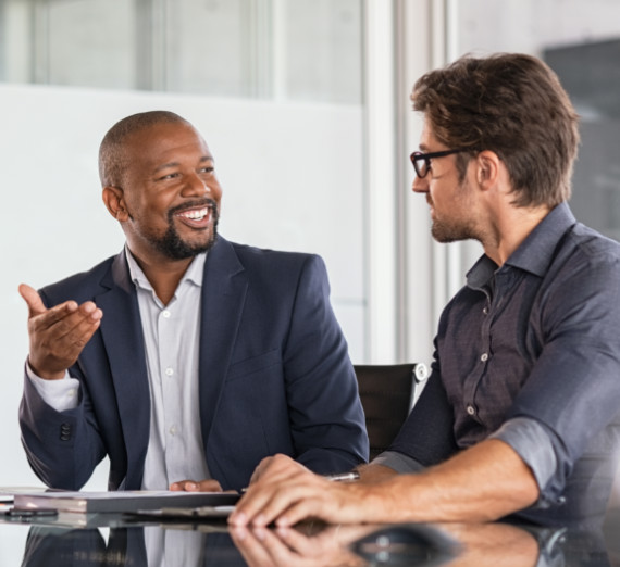 Two men in business attire sitting and talking to each other with a laptop in between them 