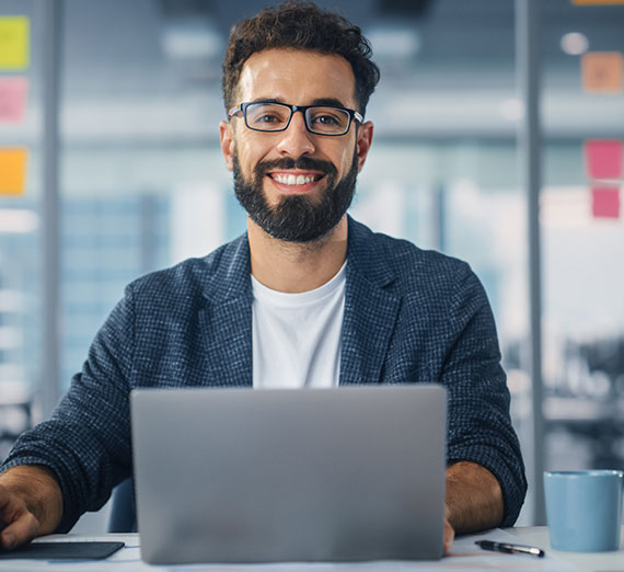 A man wearing glasses is seated in front of his laptop, smiling directly at the camera while working.