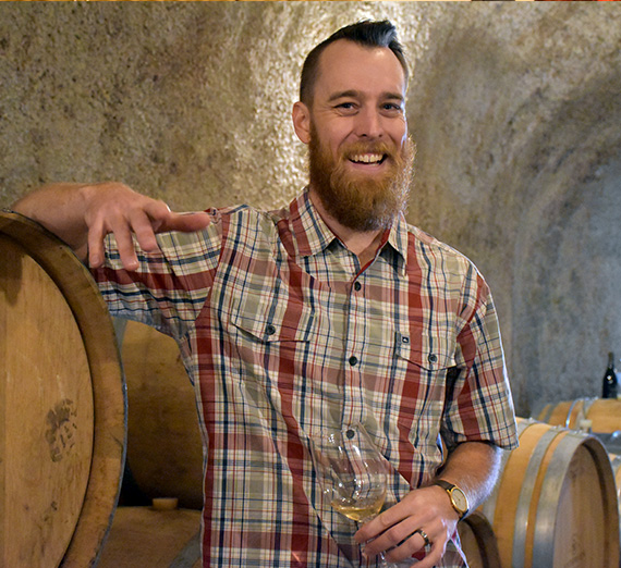 A man smiles with a glass of wine in a wine cellar.