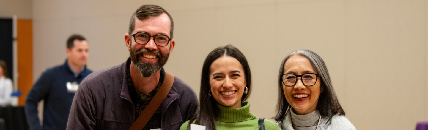 Three participants at a College of Arts & Sciences alumni networking event, including a math faculty member and two alumni panelists, standing together in an indoor venue with a neutral wall and other attendees in the background.