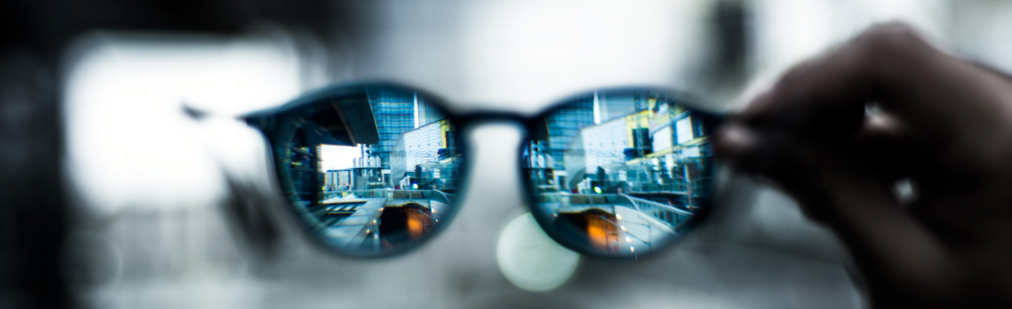 A pair of glasses being held up to view a city scene through the lenses. The image around the glasses is blurred. 