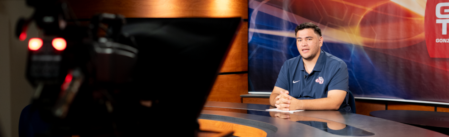Broadcast journalism student seated at a professional news anchor desk in a university television studio, with a large camera in the foreground and a colorful backdrop featuring GUTV branding.
