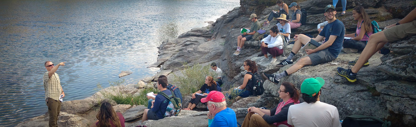 Students in the environmental studies and sciences program sit on a rock hillside near the waters edge and listen to their instructor.
