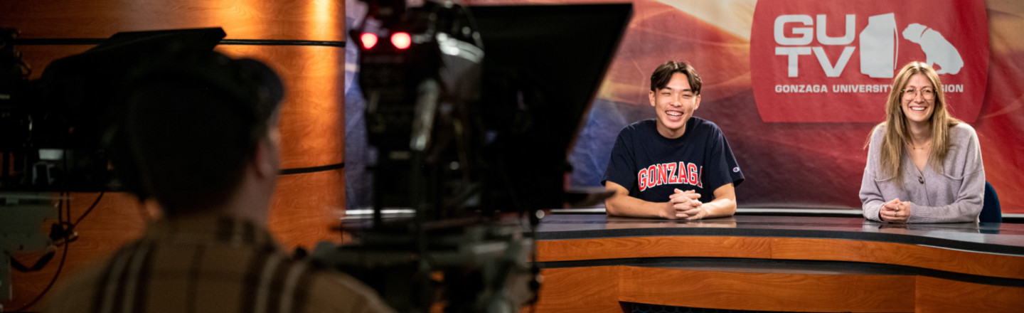 A TV studio with an out of focus camera person in the left foreground and two students in the right background at a broadcasting desk with a sign behind them reading &quot;GUTV&quot;.