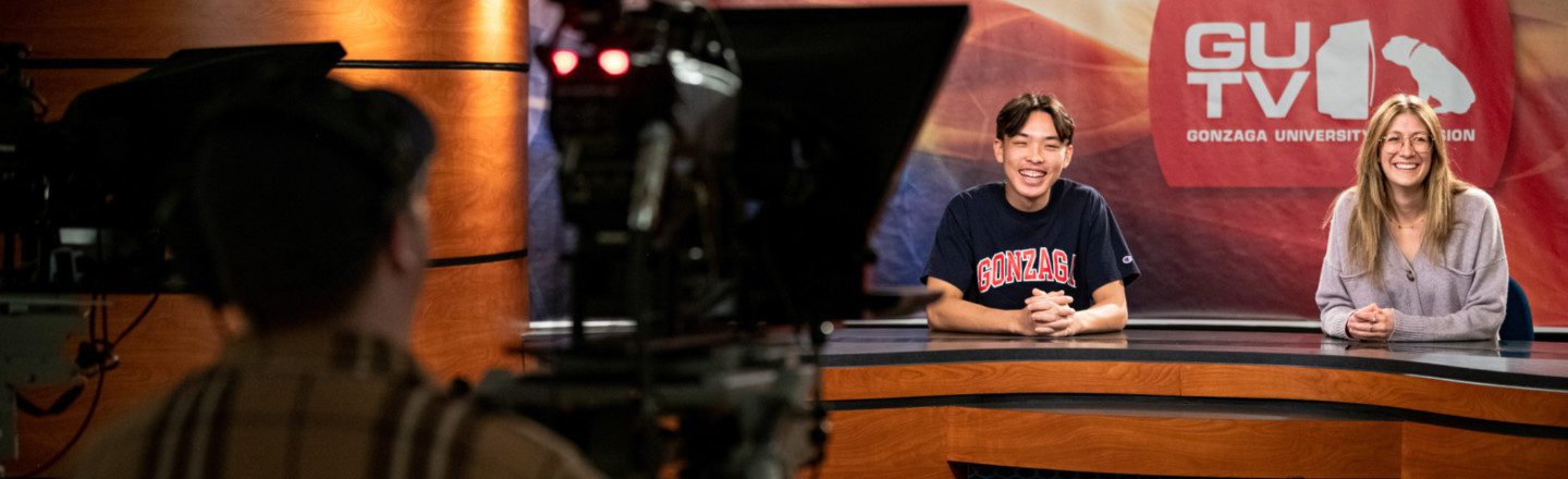 A TV studio with an out of focus camera person in the left foreground and two students in the right background at a broadcasting desk with a sign behind them reading "GUTV".