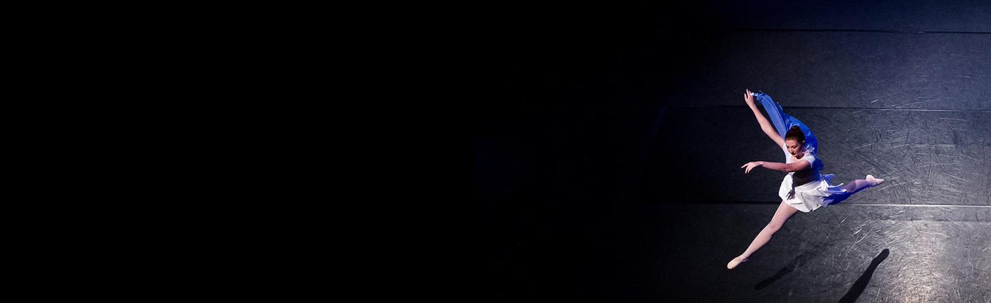 Solo dancer in white dress jumps from right to left on dark stage.