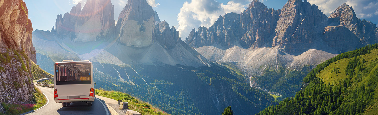A tour bus travels along a winding mountain road, surrounded by lush green hills and dramatic rocky peaks under a bright blue sky.