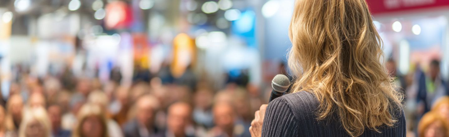 View from behind a woman with a microphone presenting to a large audience