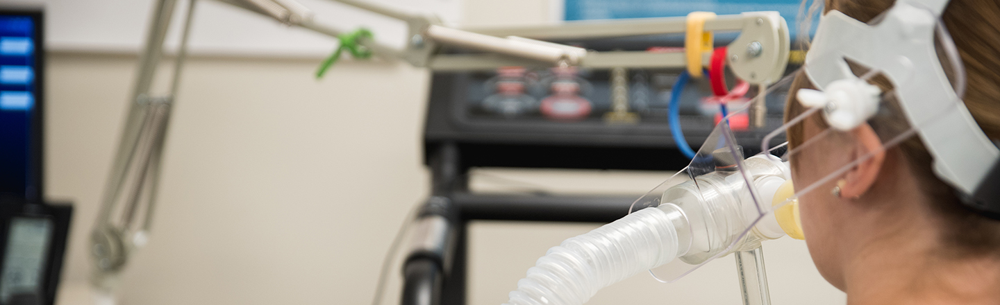  A person with their back to the camera, wearing a breathing mask and headgear, participates in a human physiology experiment. Wires and tubes from the mask connect to unseen equipment. 